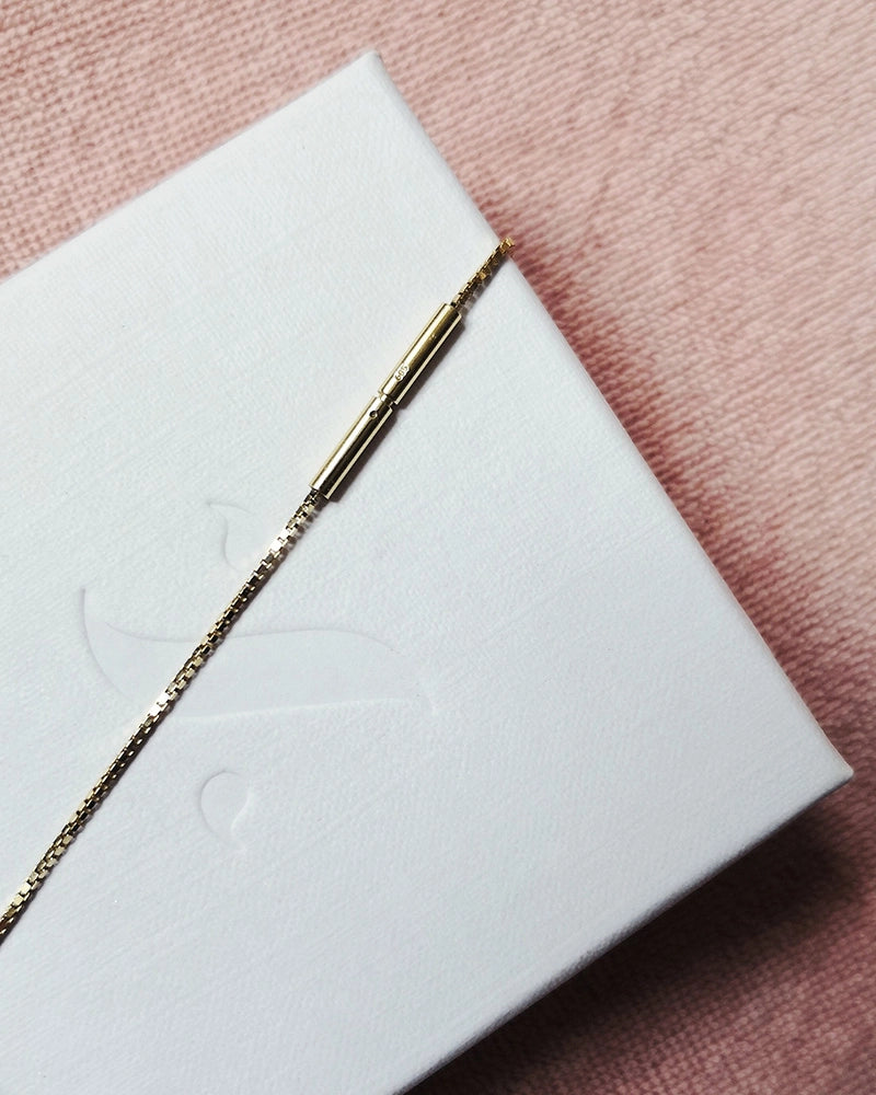 Gold memorial necklace clasp on a white box with a pink background.