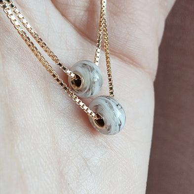 Gold memorial necklace with two gray beads on a light skin background.