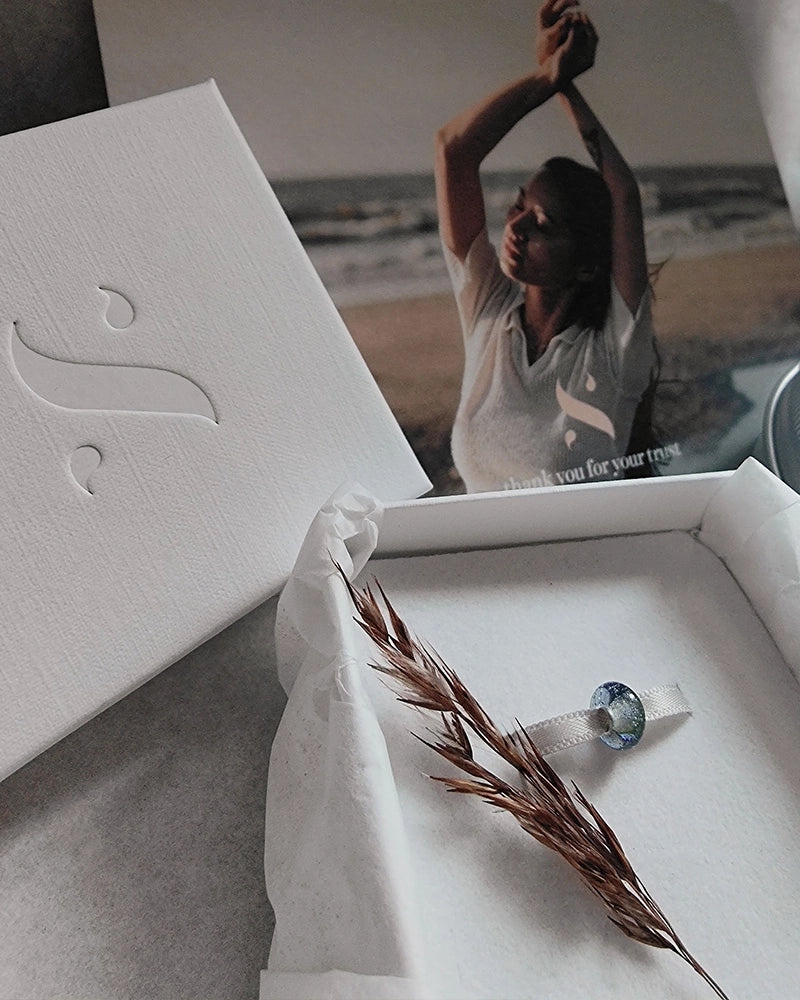 White jewelry box with a minimalist memorial pendant and dried grass, blurred background of a person with arms raised.