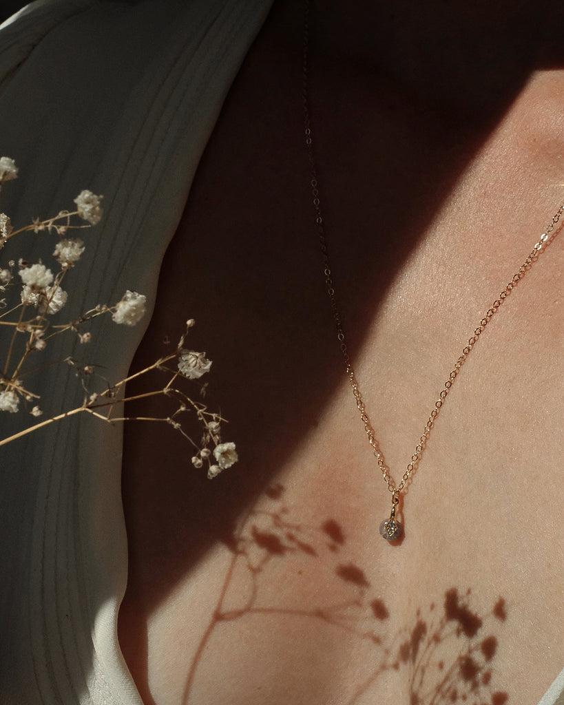 Rose gold memorial necklace with a small pendant with ashes included on a person's neck, with white flowers and shadows in the background.