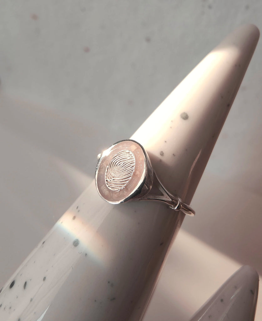 Silver cremation ring with fingerprint design on a white cylindrical stand.