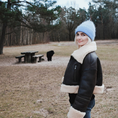 Linda Visser wearing a black jacket with white fur trim and a blue beanie standing in a park.