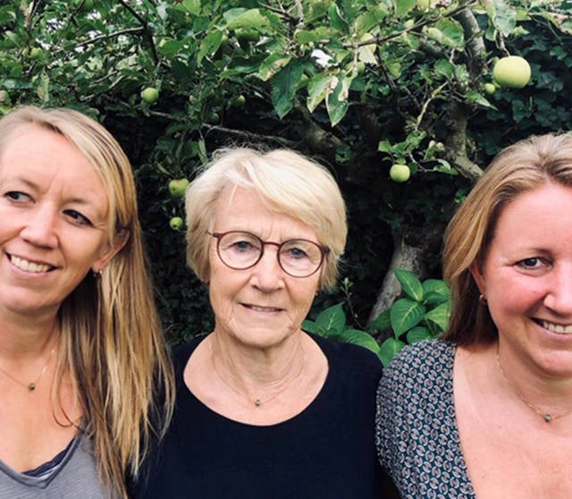 Three women standing in front of an apple tree with green apples wearing memorial jewelry.