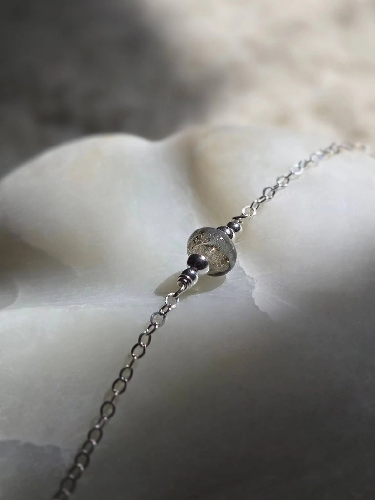 Silver memorial necklace with a cremation bead encased with ashes or hair on a textured surface.